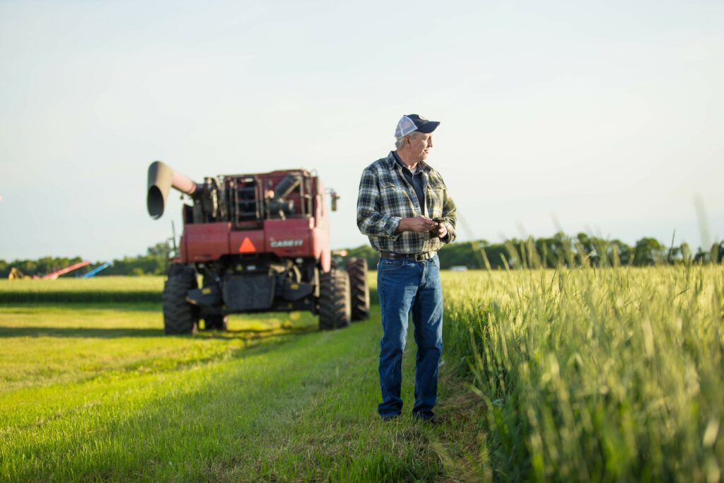 A man with a phone in his hand, looking out into the distance with a wheat field to his left and a red combine in the background