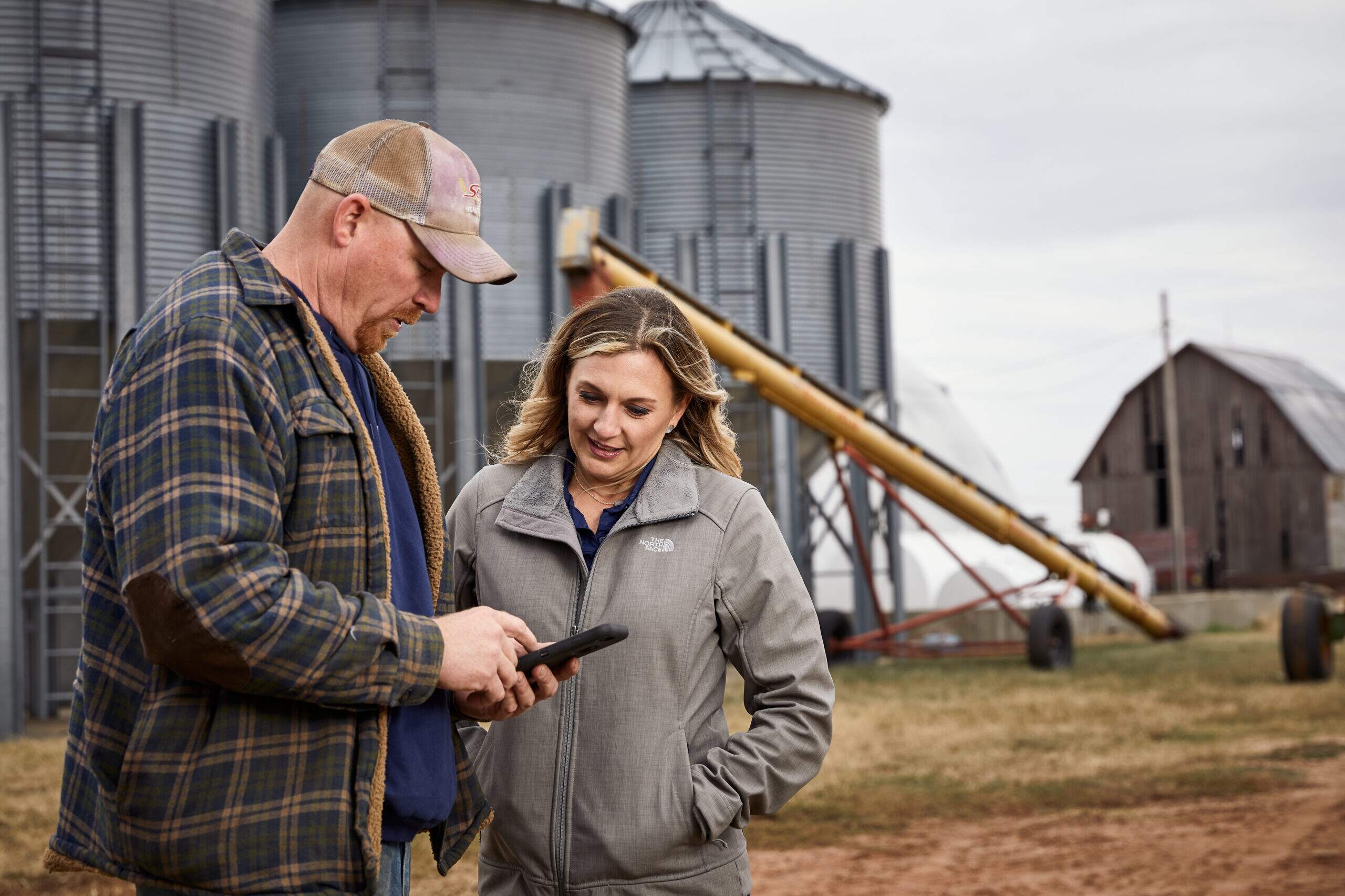 Man looks at phone with a woman looking over him as they consult on farming operations. Storage bins in the background